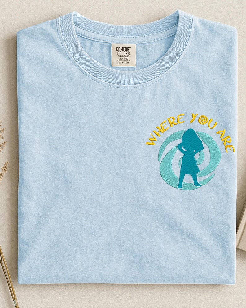 Może przedstawiać: Jasnoniebieska koszulka Comfort Colors z haftowanym napisem "WHERE YOU ARE" i turkusową sylwetką osoby. Koszulka ma okrągły dekolt i małą metkę na g&oacute;rnej lewej piersi.