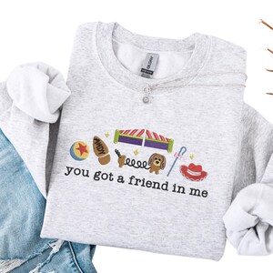 May include: A light gray sweatshirt with the embroidered phrase "you got a friend in me" and various cartoon character images. The design includes a ball, a boot, a dog, and a cowboy hat.