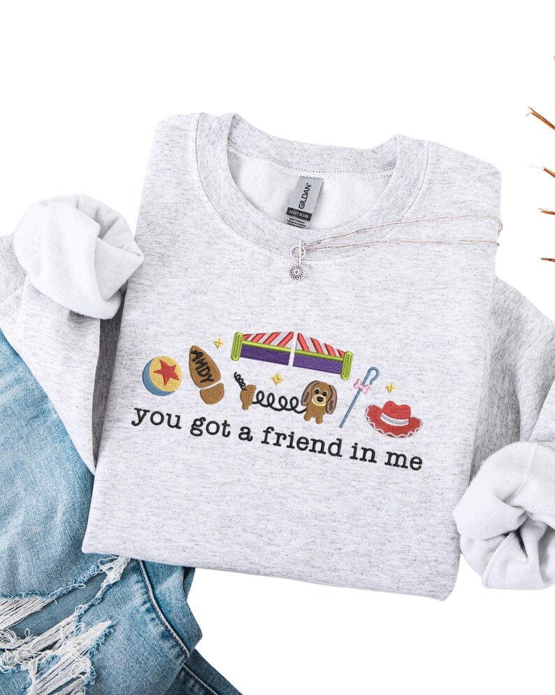 May include: A light gray sweatshirt with the embroidered phrase "you got a friend in me" and various cartoon character images. The design includes a ball, a boot, a dog, and a cowboy hat.