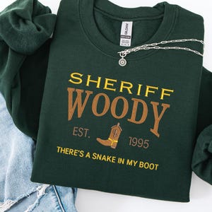 Puede incluir: Sudadera verde oscuro con el texto bordado "SHERIFF WOODY" en oro y marr&oacute;n. El dise&ntilde;o incluye una bota de vaquero y la frase "THERE'S A SNAKE IN MY BOOT". Se ve un collar plateado con un colgante de sol.