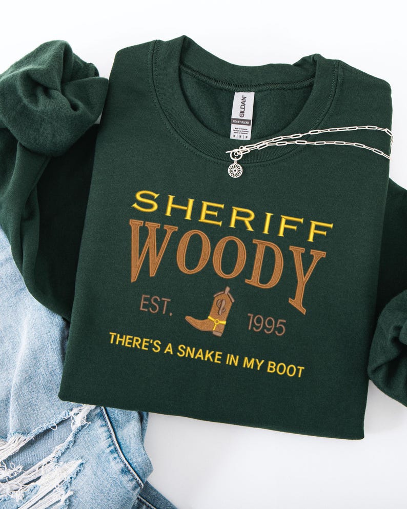 Puede incluir: Sudadera verde oscuro con el texto bordado "SHERIFF WOODY" en oro y marr&oacute;n. El dise&ntilde;o incluye una bota de vaquero y la frase "THERE'S A SNAKE IN MY BOOT". Se ve un collar plateado con un colgante de sol.