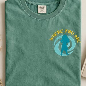 Może przedstawiać: Zielona koszulka z haftowanym napisem "WHERE YOU ARE" w kolorze ż&oacute;łtym i niebieską grafiką przedstawiającą postać w wirze. Koszulka ma okrągły dekolt i metkę "COMFORT COLORS".