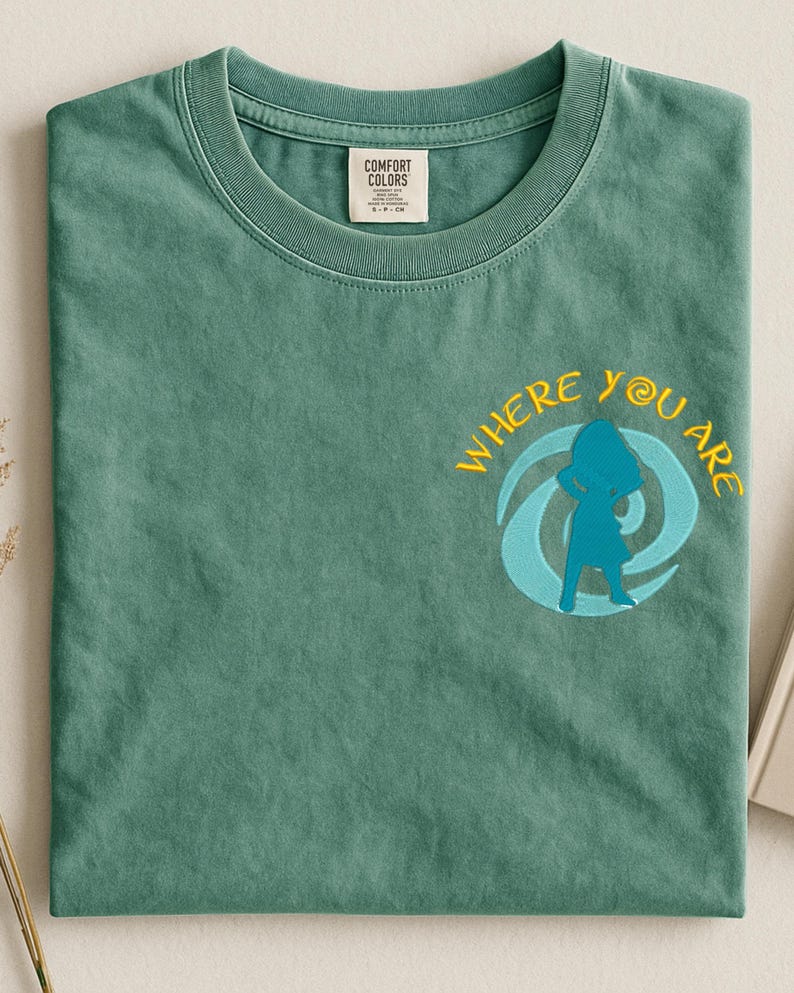 Może przedstawiać: Zielona koszulka z haftowanym napisem "WHERE YOU ARE" w kolorze ż&oacute;łtym i niebieską grafiką przedstawiającą postać w wirze. Koszulka ma okrągły dekolt i metkę "COMFORT COLORS".