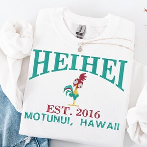 May include: White sweatshirt with the word "HEIHEI" in teal arched across the top. Below is a cartoon rooster and the text "EST. 2016 MOTUNUI, HAWAII" in teal and red. A silver necklace with a small pendant rests on the sweatshirt.