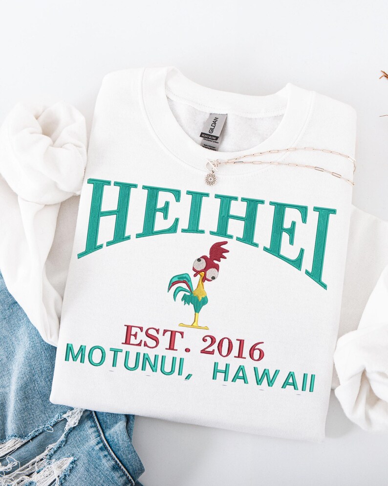 May include: White sweatshirt with the word "HEIHEI" in teal arched across the top. Below is a cartoon rooster and the text "EST. 2016 MOTUNUI, HAWAII" in teal and red. A silver necklace with a small pendant rests on the sweatshirt.