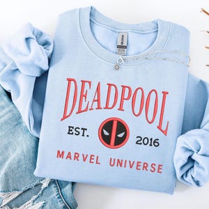 May include: A pale blue sweatshirt featuring the word "DEADPOOL" in red, with "EST. 2016 MARVEL UNIVERSE" beneath. A silver necklace with a pendant rests on the sweatshirt. The sweatshirt is folded on top of a pair of blue jeans.