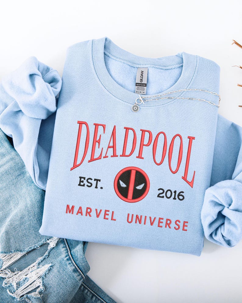 May include: A pale blue sweatshirt featuring the word "DEADPOOL" in red, with "EST. 2016 MARVEL UNIVERSE" beneath. A silver necklace with a pendant rests on the sweatshirt. The sweatshirt is folded on top of a pair of blue jeans.