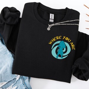 Może przedstawiać: Czarna bluza z okrągłym dekoltem z ż&oacute;łtym napisem "WHERE YOU ARE" i turkusową grafiką osoby. Srebrny naszyjnik z okrągłym wisiorkiem jest na bluzie. Bluza jest złożona na niebieskich dżinsach.