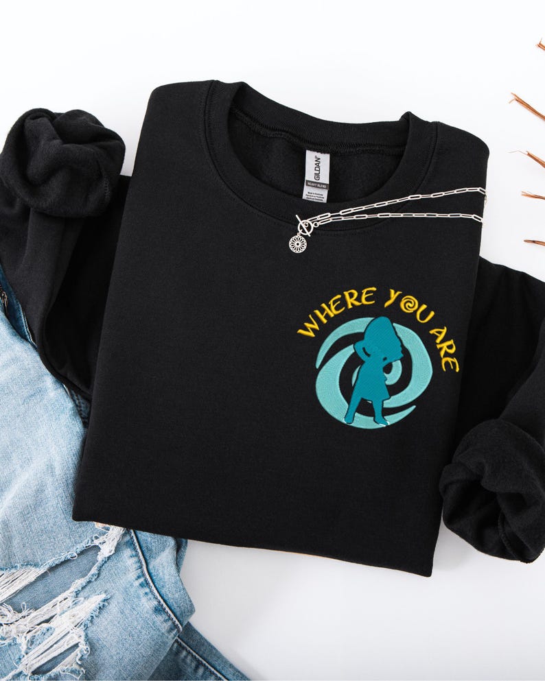 Może przedstawiać: Czarna bluza z okrągłym dekoltem z ż&oacute;łtym napisem "WHERE YOU ARE" i turkusową grafiką osoby. Srebrny naszyjnik z okrągłym wisiorkiem jest na bluzie. Bluza jest złożona na niebieskich dżinsach.