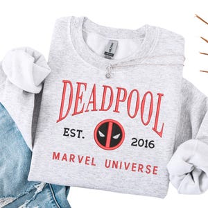 May include: A light grey sweatshirt featuring the word "DEADPOOL" in red, arched across the top. Below, the text "EST. 2016" and "MARVEL UNIVERSE" are printed in red. The Deadpool logo is in the center. A silver necklace is draped across the sweatshirt.