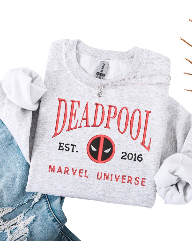 May include: A light grey sweatshirt featuring the word "DEADPOOL" in red, arched across the top. Below, the text "EST. 2016" and "MARVEL UNIVERSE" are printed in red. The Deadpool logo is in the center. A silver necklace is draped across the sweatshirt.