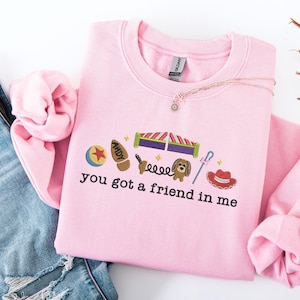 May include: A light pink sweatshirt with the phrase "you got a friend in me" and various cartoon character graphics. The sweatshirt is made of a soft material and has a crew neck.