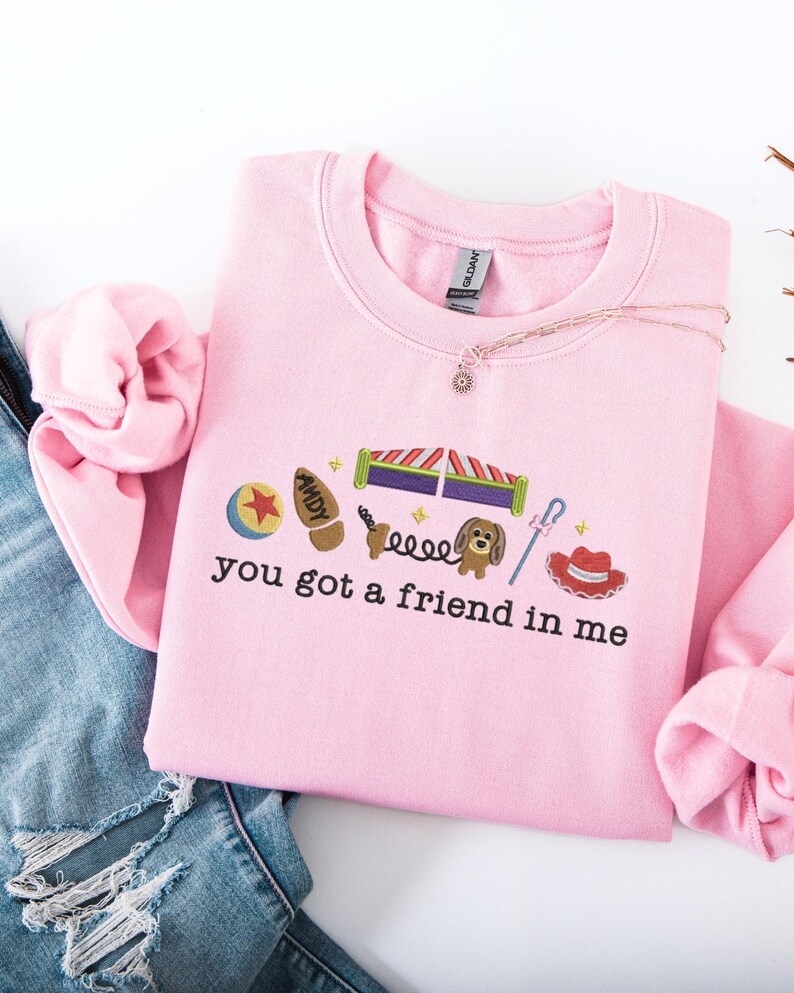 May include: A light pink sweatshirt with the phrase "you got a friend in me" and various cartoon character graphics. The sweatshirt is made of a soft material and has a crew neck.