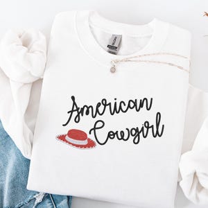May include: White sweatshirt featuring the text "American Cowgirl" in black script, with a red and white cowboy hat graphic. A silver necklace with a small pendant is on the shirt. The sweatshirt is folded on blue denim.