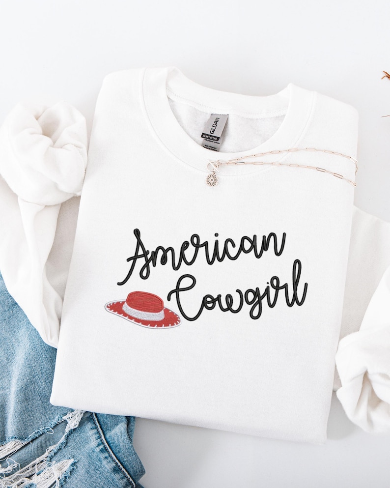 May include: White sweatshirt featuring the text "American Cowgirl" in black script, with a red and white cowboy hat graphic. A silver necklace with a small pendant is on the shirt. The sweatshirt is folded on blue denim.