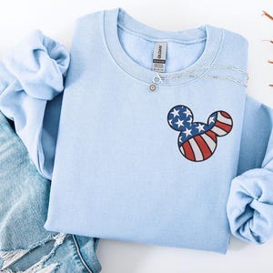 Peut inclure: Un sweat-shirt bleu clair orn&eacute; d'un motif t&ecirc;te de Mickey Mouse aux couleurs du drapeau am&eacute;ricain. Le motif comprend des rayures rouges et blanches et des &eacute;toiles bleues. Un collier argent&eacute; est drap&eacute; sur l'encolure. Le sweat-shirt est pli&eacute; sur un jean bleu.