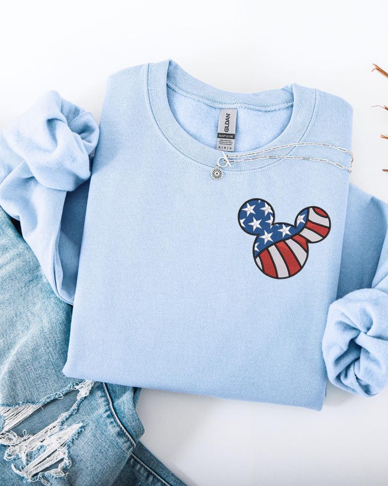 Peut inclure: Un sweat-shirt bleu clair orn&eacute; d'un motif t&ecirc;te de Mickey Mouse aux couleurs du drapeau am&eacute;ricain. Le motif comprend des rayures rouges et blanches et des &eacute;toiles bleues. Un collier argent&eacute; est drap&eacute; sur l'encolure. Le sweat-shirt est pli&eacute; sur un jean bleu.