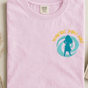 Może przedstawiać: Jasnor&oacute;żowa koszulka Comfort Colors z okrągłym dekoltem. Koszulka posiada haftowany wz&oacute;r kobiety w turkusowej sylwetce, otoczonej turkusowym i ż&oacute;łtym wirkiem, ze słowami "WHERE YOU ARE" w kolorze ż&oacute;łtym.