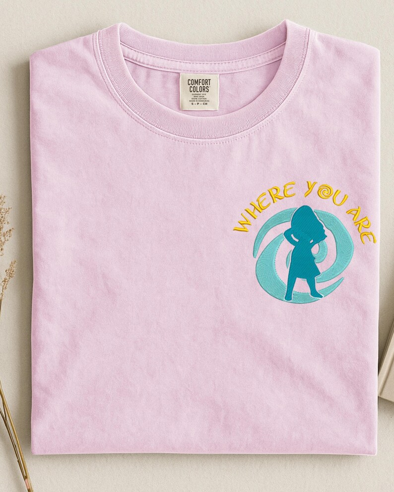 Może przedstawiać: Jasnor&oacute;żowa koszulka Comfort Colors z okrągłym dekoltem. Koszulka posiada haftowany wz&oacute;r kobiety w turkusowej sylwetce, otoczonej turkusowym i ż&oacute;łtym wirkiem, ze słowami "WHERE YOU ARE" w kolorze ż&oacute;łtym.