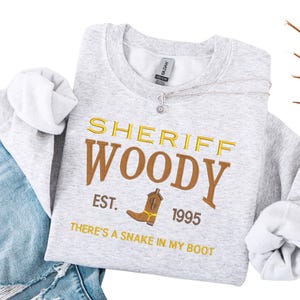 Puede incluir: Una sudadera gris claro con el texto "SHERIFF WOODY" en dorado y marr&oacute;n, junto con un dibujo de bota de vaquero. Tambi&eacute;n se ve el texto "EST. 1995" y "THERE'S A SNAKE IN MY BOOT". La sudadera est&aacute; doblada sobre unos vaqueros azules.