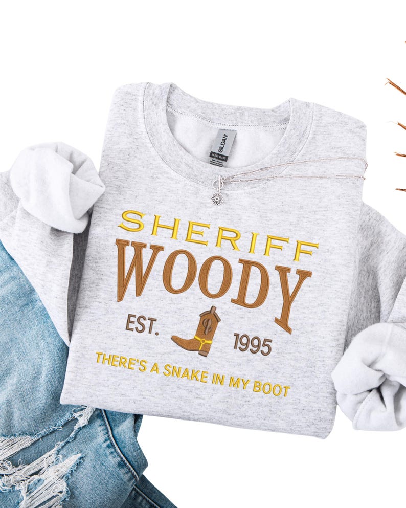 Puede incluir: Una sudadera gris claro con el texto "SHERIFF WOODY" en dorado y marr&oacute;n, junto con un dibujo de bota de vaquero. Tambi&eacute;n se ve el texto "EST. 1995" y "THERE'S A SNAKE IN MY BOOT". La sudadera est&aacute; doblada sobre unos vaqueros azules.