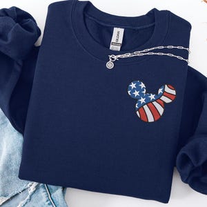 Peut inclure: Sweat-shirt bleu marine avec un motif Mickey Mouse aux couleurs du drapeau am&eacute;ricain. Un collier argent&eacute; avec un pendentif soleil est drap&eacute; sur le sweat-shirt. Le sweat-shirt est pli&eacute; sur un jean bleu.