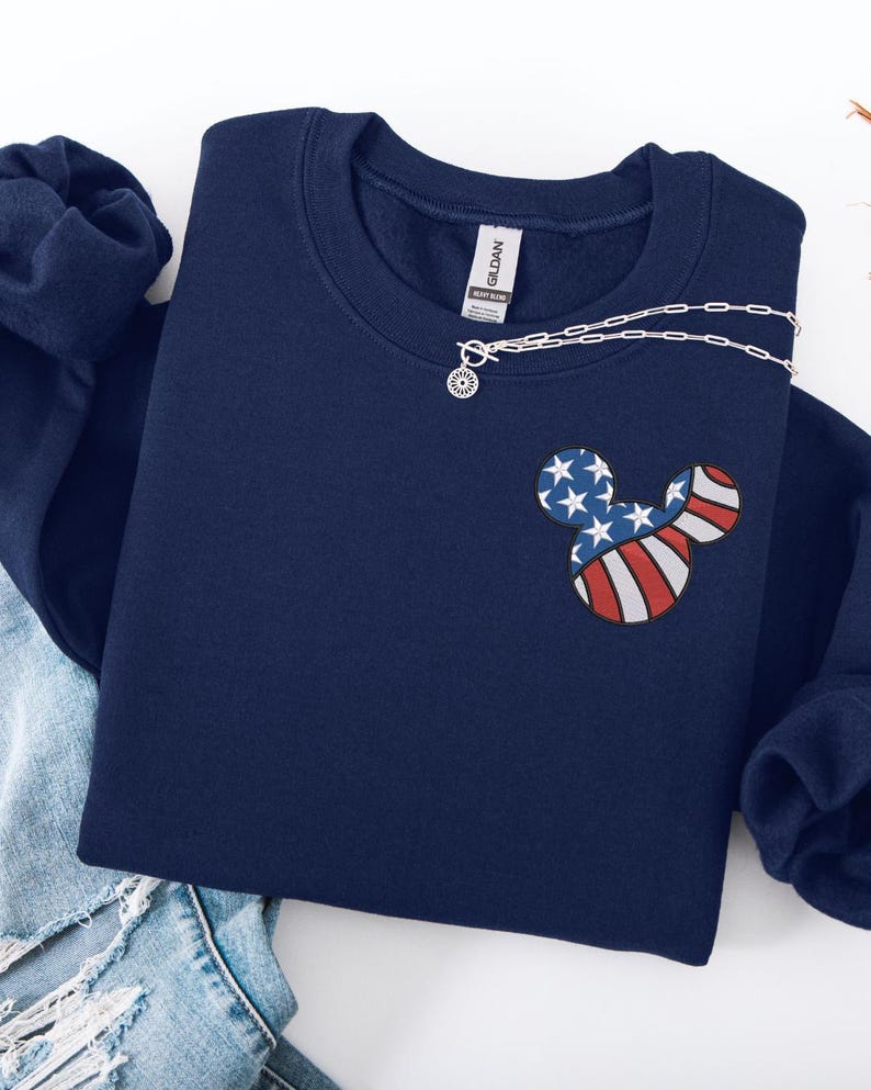 Peut inclure: Sweat-shirt bleu marine avec un motif Mickey Mouse aux couleurs du drapeau am&eacute;ricain. Un collier argent&eacute; avec un pendentif soleil est drap&eacute; sur le sweat-shirt. Le sweat-shirt est pli&eacute; sur un jean bleu.