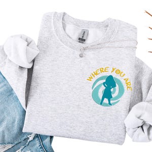 Może przedstawiać: Jasnoszara bluza z napisem "WHERE YOU ARE" w kolorze ż&oacute;łtym i turkusową grafiką. Widoczny srebrny naszyjnik z zawieszką w kształcie kwiatu. Bluza jest połączona z niebieskimi dżinsami.