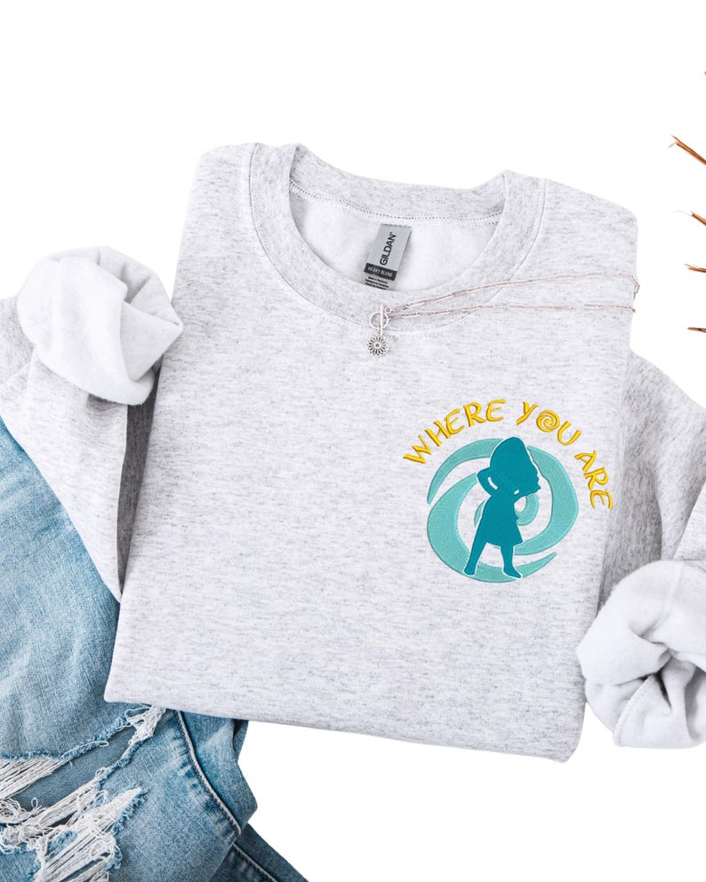 Może przedstawiać: Jasnoszara bluza z napisem "WHERE YOU ARE" w kolorze ż&oacute;łtym i turkusową grafiką. Widoczny srebrny naszyjnik z zawieszką w kształcie kwiatu. Bluza jest połączona z niebieskimi dżinsami.