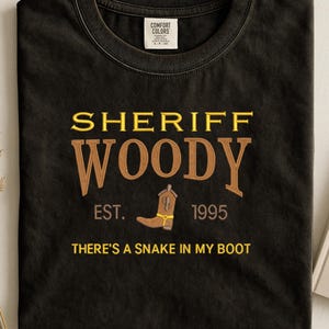 Puede incluir: Camiseta negra con las palabras "SHERIFF WOODY" en letras doradas y marrones. Incluye una bota de vaquero marr&oacute;n y la frase "THERE'S A SNAKE IN MY BOOT". La camiseta tambi&eacute;n tiene el texto "EST. 1995".