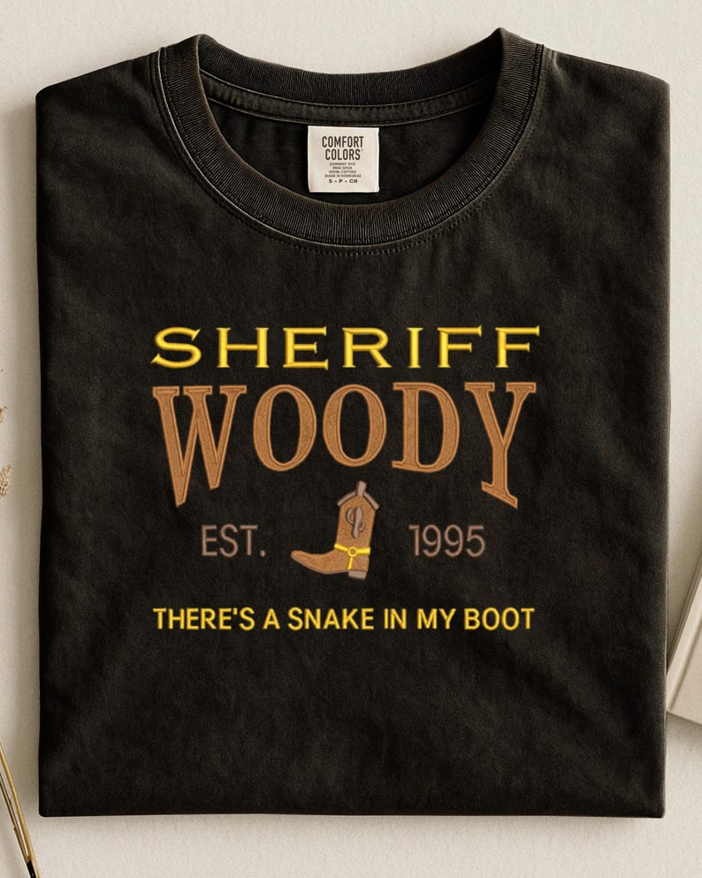 Puede incluir: Camiseta negra con las palabras "SHERIFF WOODY" en letras doradas y marrones. Incluye una bota de vaquero marr&oacute;n y la frase "THERE'S A SNAKE IN MY BOOT". La camiseta tambi&eacute;n tiene el texto "EST. 1995".