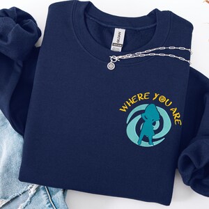 Może przedstawiać: Granatowa bluza z napisem "WHERE YOU ARE" i turkusową grafiką. Srebrny naszyjnik z zawieszką w kształcie słońca spoczywa na bluzie. Bluza jest złożona na parze niebieskich dżins&oacute;w.