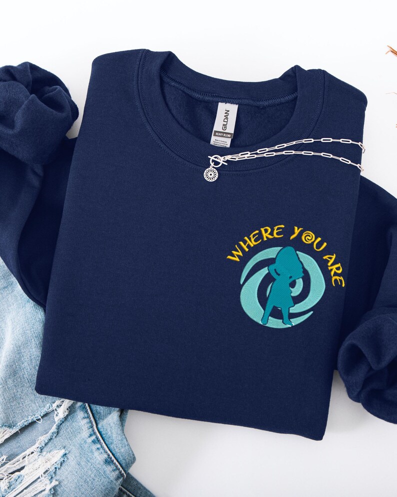 Może przedstawiać: Granatowa bluza z napisem "WHERE YOU ARE" i turkusową grafiką. Srebrny naszyjnik z zawieszką w kształcie słońca spoczywa na bluzie. Bluza jest złożona na parze niebieskich dżins&oacute;w.