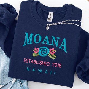K&ouml;nnte beinhalten: Ein marineblaues Sweatshirt mit dem in T&uuml;rkis gestickten Wort "MOANA", einem t&uuml;rkisfarbenen Wirbeldesign und rosa Hibiskusbl&uuml;ten. Der Text "ESTABLISHED 2016 HAWAII" steht darunter. Eine silberne Halskette mit Sonnenanh&auml;nger liegt darauf.