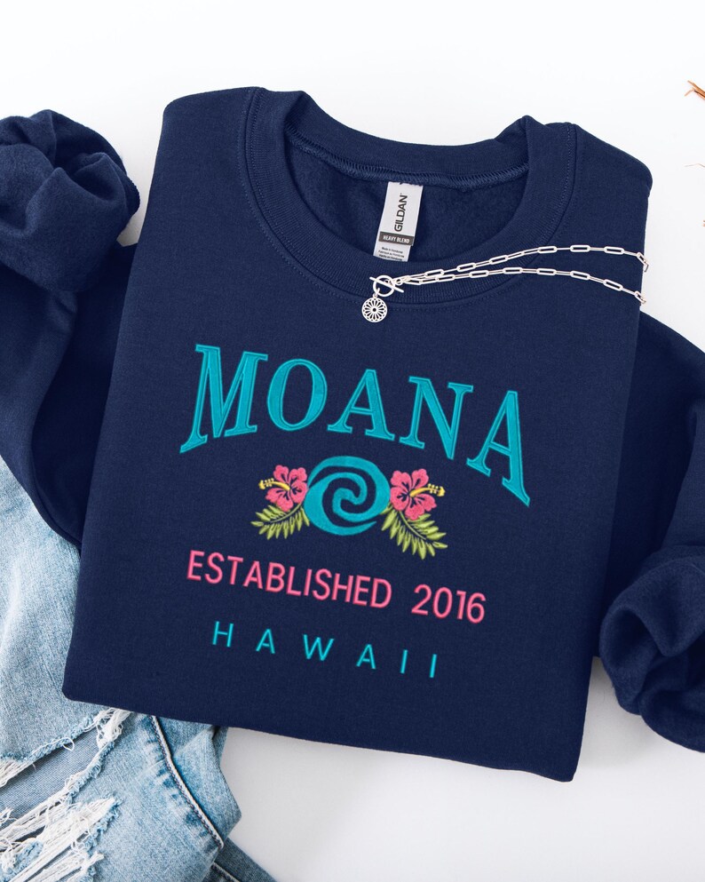 K&ouml;nnte beinhalten: Ein marineblaues Sweatshirt mit dem in T&uuml;rkis gestickten Wort "MOANA", einem t&uuml;rkisfarbenen Wirbeldesign und rosa Hibiskusbl&uuml;ten. Der Text "ESTABLISHED 2016 HAWAII" steht darunter. Eine silberne Halskette mit Sonnenanh&auml;nger liegt darauf.