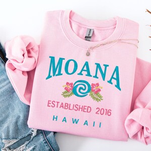 K&ouml;nnte beinhalten: Ein hellrosa Sweatshirt mit dem Wort "MOANA" in T&uuml;rkis gestickt, zusammen mit einem Blumendesign und dem Text "ESTABLISHED 2016 HAWAII". Eine goldene Halskette mit Sonnenanh&auml;nger liegt auf dem Sweatshirt. Das Sweatshirt ist neben einer blauen Jeans.