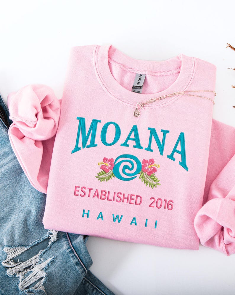 K&ouml;nnte beinhalten: Ein hellrosa Sweatshirt mit dem Wort "MOANA" in T&uuml;rkis gestickt, zusammen mit einem Blumendesign und dem Text "ESTABLISHED 2016 HAWAII". Eine goldene Halskette mit Sonnenanh&auml;nger liegt auf dem Sweatshirt. Das Sweatshirt ist neben einer blauen Jeans.