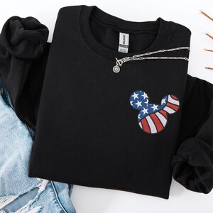 Peut inclure: Sweat-shirt noir &agrave; col rond avec un motif inspir&eacute; de Mickey Mouse aux couleurs du drapeau am&eacute;ricain. Le motif comprend des &eacute;toiles bleues et des rayures rouges et blanches. Un collier argent&eacute; avec un petit pendentif est pos&eacute; sur le sweat-shirt.
