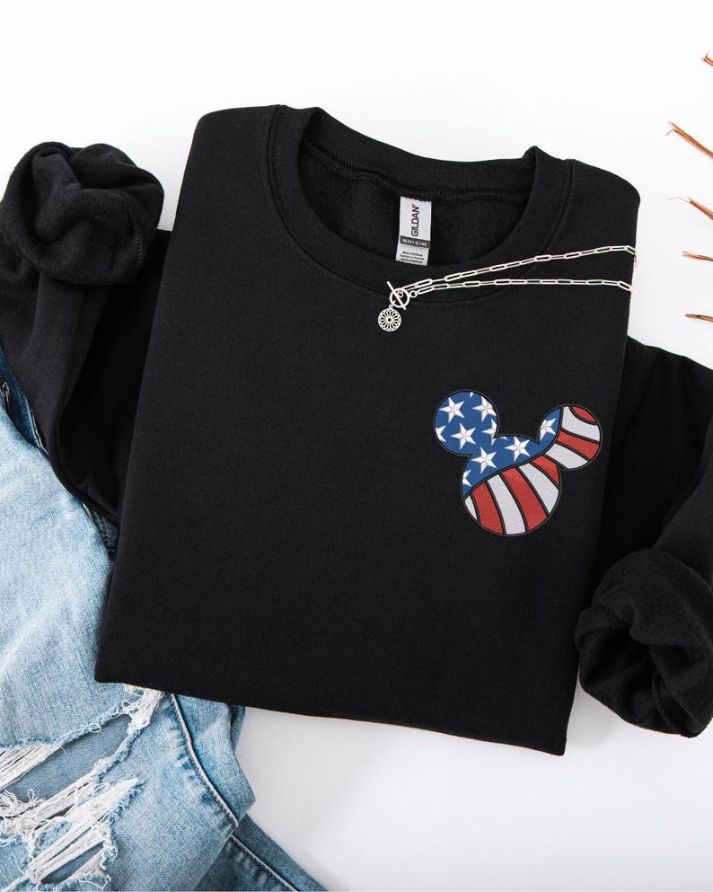 Peut inclure: Sweat-shirt noir &agrave; col rond avec un motif inspir&eacute; de Mickey Mouse aux couleurs du drapeau am&eacute;ricain. Le motif comprend des &eacute;toiles bleues et des rayures rouges et blanches. Un collier argent&eacute; avec un petit pendentif est pos&eacute; sur le sweat-shirt.