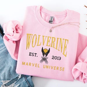 May include: A pale pink sweatshirt featuring the word "WOLVERINE" in gold. Below is a black and gold graphic of Wolverine's mask and claws, with "EST. 2013 MARVEL UNIVERSE". A gold necklace with a small pendant is draped across the sweatshirt.