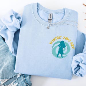 Może przedstawiać: Jasnoniebieska bluza z napisem "WHERE YOU ARE" w kolorze ż&oacute;łtym i turkusową sylwetką osoby. Srebrny naszyjnik z zawieszką w kształcie słońca leży na bluzie. Bluza jest złożona na parze dżins&oacute;w.