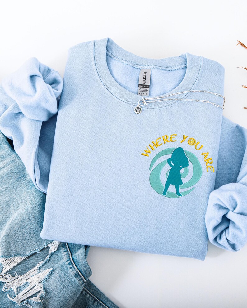 Może przedstawiać: Jasnoniebieska bluza z napisem "WHERE YOU ARE" w kolorze ż&oacute;łtym i turkusową sylwetką osoby. Srebrny naszyjnik z zawieszką w kształcie słońca leży na bluzie. Bluza jest złożona na parze dżins&oacute;w.