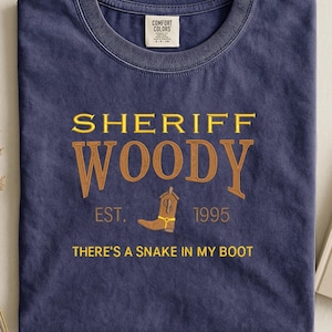 Puede incluir: Camiseta azul marino con el texto "SHERIFF WOODY" en letras doradas y marrones. La camiseta presenta una bota de vaquero y la frase "THERE'S A SNAKE IN MY BOOT". La camiseta es de 1995.