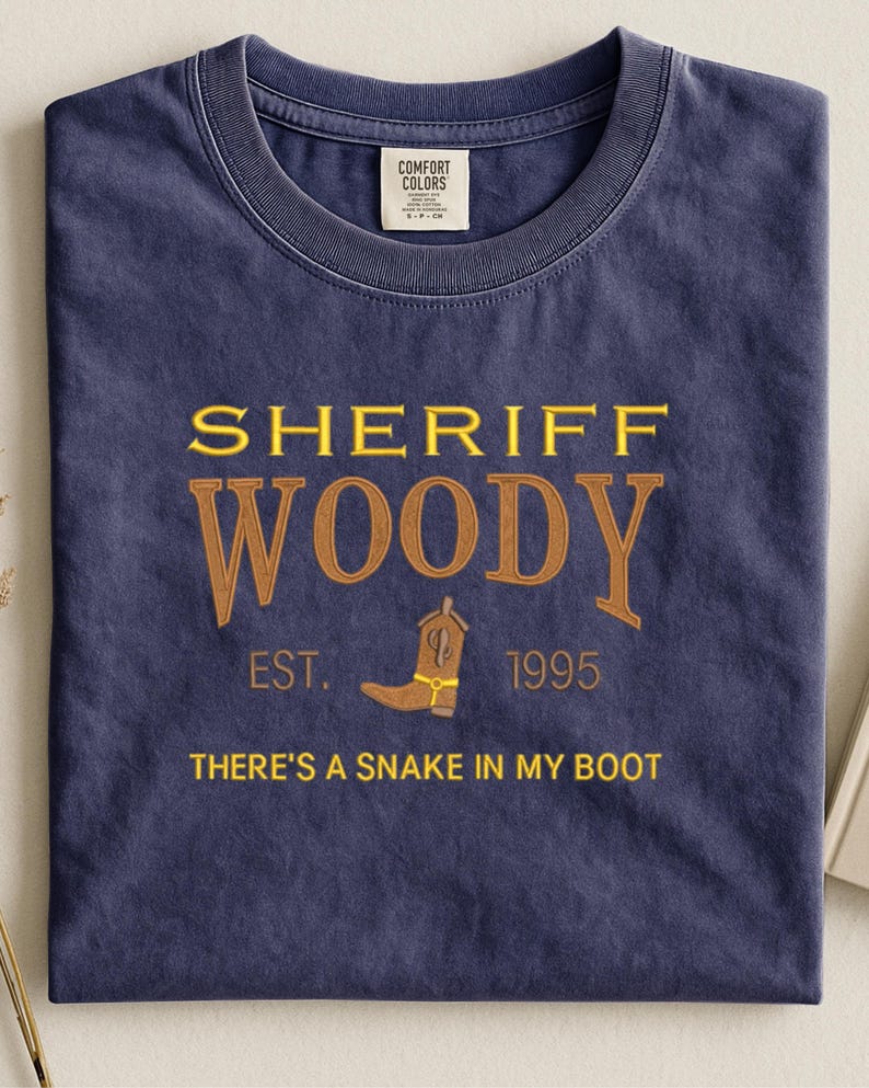 Puede incluir: Camiseta azul marino con el texto "SHERIFF WOODY" en letras doradas y marrones. La camiseta presenta una bota de vaquero y la frase "THERE'S A SNAKE IN MY BOOT". La camiseta es de 1995.