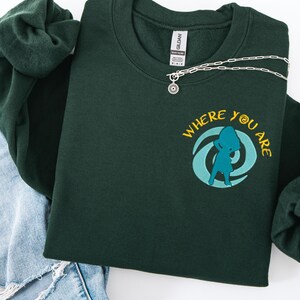 Może przedstawiać: Ciemnozielona bluza z napisem "WHERE YOU ARE" i turkusową grafiką. Srebrny naszyjnik z okrągłą zawieszką leży na bluzie. Bluza jest złożona na parze niebieskich dżins&oacute;w i ciemnozielonej gumce do włos&oacute;w.