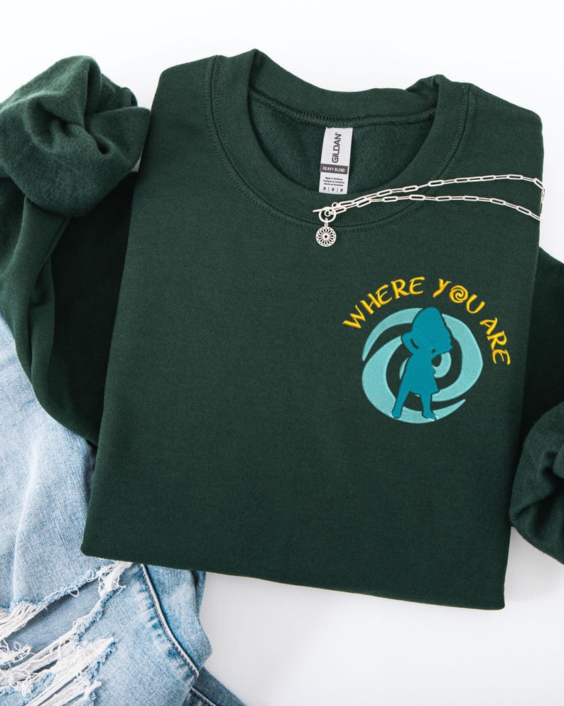 Może przedstawiać: Ciemnozielona bluza z napisem "WHERE YOU ARE" i turkusową grafiką. Srebrny naszyjnik z okrągłą zawieszką leży na bluzie. Bluza jest złożona na parze niebieskich dżins&oacute;w i ciemnozielonej gumce do włos&oacute;w.