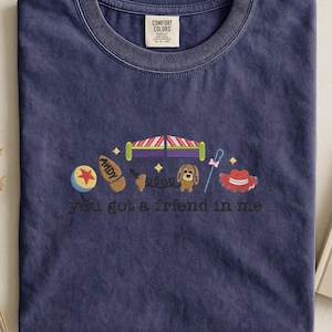 May include: Navy blue t-shirt with embroidered cartoon images and the text "you got a friend in me." The design includes a ball, a shoe, a bed, a dog, a spring, a hat, and a cane. The shirt has a crew neck and a Comfort Colors tag.