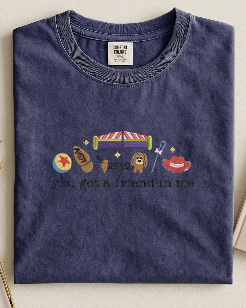 May include: Navy blue t-shirt with embroidered cartoon images and the text "you got a friend in me." The design includes a ball, a shoe, a bed, a dog, a spring, a hat, and a cane. The shirt has a crew neck and a Comfort Colors tag.