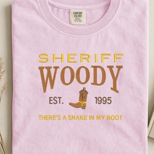 Puede incluir: Camiseta rosa claro con bordado dorado y marr&oacute;n. El texto "SHERIFF WOODY" est&aacute; bordado, con una bota de vaquero marr&oacute;n y la frase "THERE'S A SNAKE IN MY BOOT" debajo. La camiseta tambi&eacute;n tiene el texto "EST. 1995".