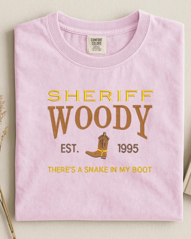 Puede incluir: Camiseta rosa claro con bordado dorado y marr&oacute;n. El texto "SHERIFF WOODY" est&aacute; bordado, con una bota de vaquero marr&oacute;n y la frase "THERE'S A SNAKE IN MY BOOT" debajo. La camiseta tambi&eacute;n tiene el texto "EST. 1995".